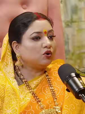 The image is a close-up of a woman speaking into a microphone. She is wearing a yellow outfit, ornate gold jewelry, and has a red bindi on her forehead. The background is blurred, suggesting she may be in a studio or event setting.