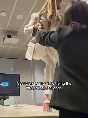 A woman in a white suit is standing on a table, supported in part by a second person. The hook text "Nearly broke my ankle trying the Nikki Minaj challenge" is overlaid at the bottom of the screen. The background is an office.