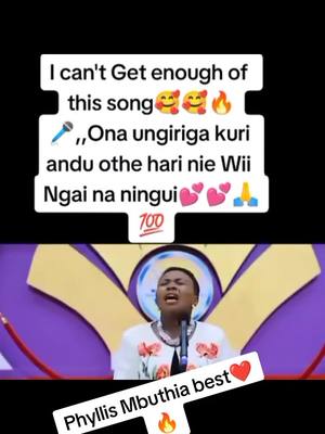 The image shows a woman singing into a microphone, likely in a church or studio setting. She's wearing a white top with a necklace. Text overlays include a song recommendation and the name "Phyllis Mbuthia best". Background features purple and gold decorative elements. A microphone graphic and emojis are present in the overlay text highlighting the song.