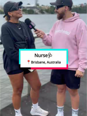 A cover image of a man and a woman standing by the water. The woman in the foreground is holding a microphone, and the man is holding the microphone as well. The woman is wearing a black cap, sunglasses, a black sweatshirt-style top, and black shorts. The man is wearing a black cap, sunglasses, and a pink hoodie. A text box at the bottom indicates that the video will be based in Brisbane, Australia and will focus on nurses.