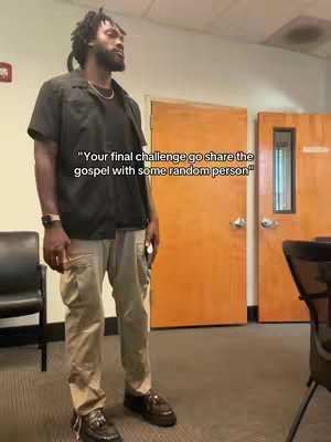 A man stands in a brightly lit room. He wears a black collared shirt over a black t-shirt, paired with light beige cargo pants and dark brown shoes, with what appears to be dreadlocks. In the background are closed doors with wooden paneling, and an empty chair. Text overlay stating Your final challenge go share the gospel with some random person.
