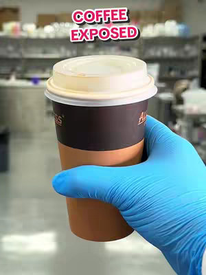 A hand wearing a blue glove holds a coffee cup. The cup is brown with a white lid. The background is blurry, but appears to be a lab setting. Text overlay says 'COFFEE EXPOSED'.