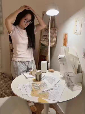 The image shows a young woman in a softly lit room, standing near a round white table. She is wearing a white t-shirt and striped shorts, her long dark hair cascading down. Her hands are raised towards her head, as if frustrated. On the table is a spilled coffee stain over papers indicative of a chaotic moment. A floor lamp adds a warm glow, while a mirror, a decorative bear figure, and other items adorn the background, creating a cozy and lived-in atmosphere.