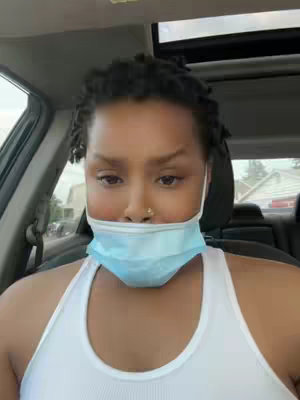 A selfie taken from inside a car shows a woman with a mask pulled down below her nose. She has dark hair in small twists. She wears a white tank top, and the car’s interior is visible in the background.