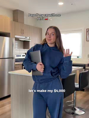 A young woman in a blue sweatsuit holds a laptop and waves in a kitchen setting. Text overlays read 'Payoff my car with me Pt. 3' and 'let's make my $4,000.' The background features a kitchen with wooden cabinets, stainless steel appliances, and a breakfast bar. The lighting is bright and natural.