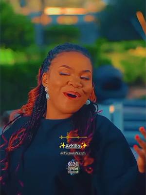 The image is a medium shot of a woman with dark skin, her eyes closed and mouth slightly open as if she's singing or speaking passionately. Her hair is styled with long braids, some of which are dyed a reddish-orange color. She wears large, silver earrings. Behind her, the background is blurred, with hints of green foliage and a light-colored structure, possibly a building or wall. Overlayed on the image are text elements.