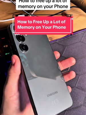 A person holds a gray Samsung phone, angled to show the back, with a red text box overlay at the center. The background is a desk setting with tech-related items.