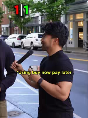 A man holding a microphone interviews someone, with a busy street scene in the background that includes cars and buildings.