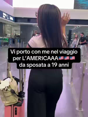 A woman is shown from behind, standing in what appears to be an airport terminal. She's wearing glasses, a white top and black pants, and is holding her hand up. There is text overlaid on the image in a banner towards the bottom.