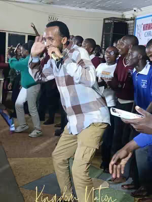 A man with a microphone is in the foreground, mid-action. He has a checkered shirt and khaki pants, and is gesturing with his hand. Behind him, a crowd of young people are gathered in what appears to be an event or meeting, some raising their hands. The lighting is warm, and the background is slightly blurred, highlighting the central figure. The words 'Kingdom Nation' are subtly present in the bottom of the image.