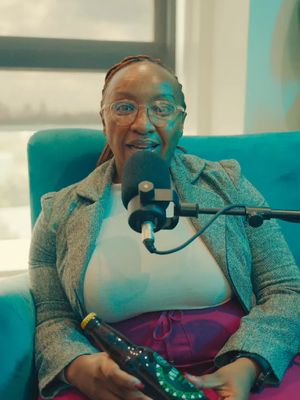 A woman with glasses is seated in front of a microphone, holding a beer bottle. The background consists of a window and a blue armchair, suggesting an interview setup.
