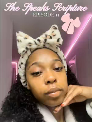 A selfie-style shot features a woman with dark, curly hair. She wears a fluffy bow headband and a white robe, and is posing with her hand under her chin in a slight pose. The background is a faint pink-toned wall.