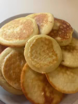 A close-up shot of a plate piled with golden-brown pancakes. They appear fluffy with slightly crisp edges, stacked and overlapping. The lighting is soft, highlighting the textures.