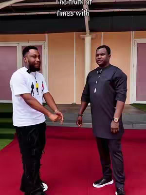 The image shows two men standing and facing each other in an outdoor setting. Both are wearing microphones. The man on the left is younger, wearing a white graphic tee, black pants, and has a styled goatee. The man on the right is older, dressed in a dark long-sleeved shirt, and appears to be in a formal outfit. The floor is covered with a red carpet, and the background includes large cream-colored pillars and walls, likely from a building facade.