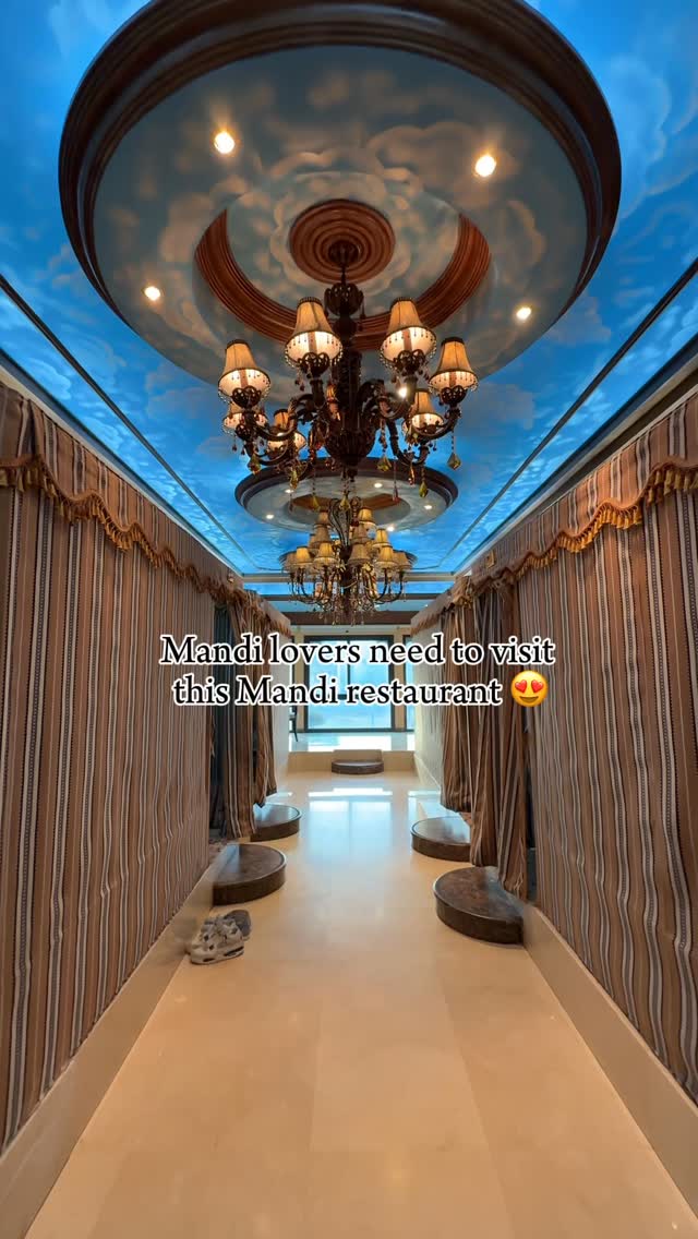 The cover image shows a hallway with ornate ceilings and walls. The ceiling is painted to resemble a blue sky with clouds, and it is adorned with a large, elaborate chandelier. Along both sides of the hallway, there are draped curtains. The floor is a light color and appears to be reflective. The perspective draws you down the corridor towards a bright, possibly windowed spot at the end. At the bottom, there is a pair of shoes on the floor, hinting that this is a real location.