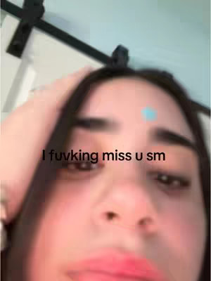 A close-up shot featuring a young woman with a slightly blurred background of a room. She has dark hair, and her eyes are focused towards the camera. A small dot appears on her forehead. The overlayed text reads: "I fuvking miss u sm".