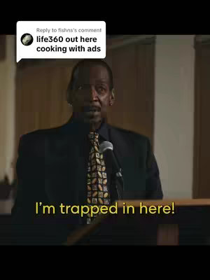 The image shows a middle-aged black man in a dark suit, tie, standing behind a podium with a microphone. In the background, there is a simple interior setting.