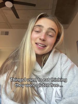 A young woman with long blonde hair is centered in the frame, wearing a gray hoodie. The top of a ceiling fan is visible in the background. Text overlayed on the image reads, "Things I used to eat thinking they were gluten free ..."