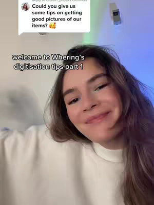 A selfie-style video with a young woman at the center, smiling. The background is a simple wall with an obscured object hanging on it, and a comment bubble. Text overlay presents "welcome to Whering's digitisation tips part 1."