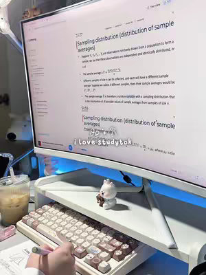 A desk setup with a computer monitor displaying a document about 'Sampling distribution'. A keyboard, a drink, and a small bear figurine are also present on the desk. The image is lit with warm, natural tones and a cool-toned blue light under the monitor stand.