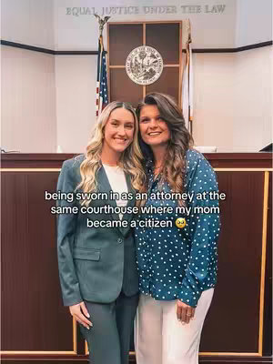 Two women are posing in a courtroom. The woman on left with blonde hair and a suit. She smiles towards the camera. The woman on the right with brown hair and a blue dotted shirt poses next to her. The background includes the American flag, the seal of the state, and a wooden paneled interior.