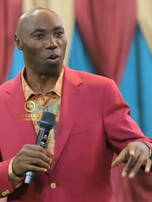A medium shot features a man with a bald head wearing a red suit, holding a microphone and speaking. The background is blurred, suggesting a stage or event setting. The man has a serious expression, and the lighting appears to be natural, illuminating his face.