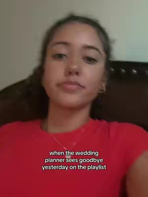 A young woman is shown in a close-up shot, appearing from the chest up against a soft, blurred background. She wears a red shirt and a delicate necklace and has dark hair. Text overlayed at the bottom of the image reads 'when the wedding planner sees goodbye yesterday on the playlist.'