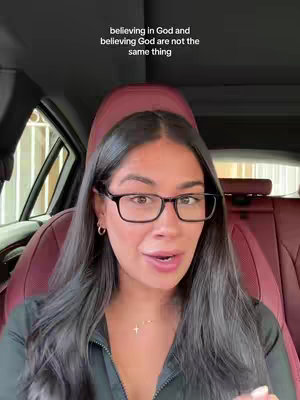 A woman with long dark hair and glasses is sitting in the driver's seat of a car. The background includes the car's headrest and window, while the woman is facing the camera and talking. Text overlay indicates that beliefs in God are not the same.