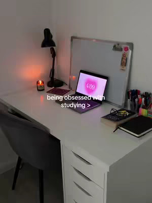 A well-lit desk with a white surface and a grey chair. A laptop displaying a pink clock, a notebook, and a book sit on the desk. A desk lamp and a candle are placed on the desk. A whiteboard and colored pens are also visible.