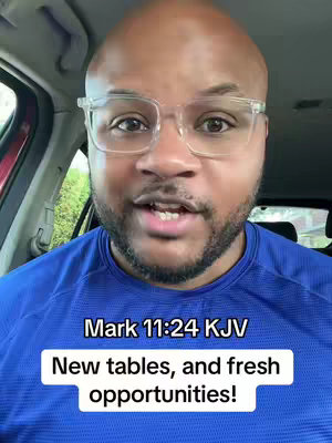 A bald Black man with a beard and glasses is positioned inside of a car, the interior visible in the background. The man is wearing a blue shirt and speaking to the camera. There is a text overlay on the bottom of the image that shows Mark 11:24 KJV and "New tables, and fresh opportunities!" 