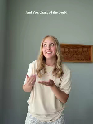 A young woman with blonde hair stands in front of a light gray wall, looking upwards with a wide, smiling expression. She wears a light beige t-shirt and patterned pants. Her arms are raised, with palms facing upward. Above her is the text overlay: 'And You changed the world', and to the right is a framed wooden relief.