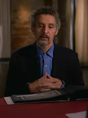 A medium shot of John Turturro sitting in front of a red table, with his hands clasped in front of him. He is wearing a dark blazer over a light blue collared shirt. Beige curtains are visible in the background.