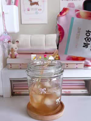 A well-lit, aesthetically pleasing shot of a study desk with a jar of tea with ice cubes in the center. Surrounding this are various stationery items and mini decorations, indicative of a study space. A cute My Melody plush toy sits on the left, and a poster of a dog is on the top. The overall composition is designed to create a cozy and appealing study atmosphere.