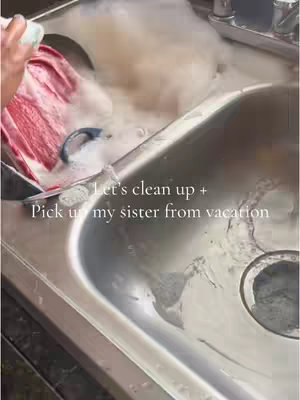 The image shows a stainless steel sink filling up with sudsy water. In the sink is a red and white dish. The text overlay says, "Let's clean up + Pick up my sister from vacation."