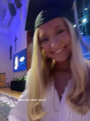 A smiling young woman in a graduation cap and gown stands in a hallway, likely at her graduation ceremony. The background includes interior architectural elements typical of a school such as doors, lighting fixtures and a large screen with graphics.