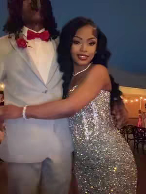 Image shows a couple at prom. The woman is in a sparkling silver gown, while the man is in a grey suit and red bow tie. They are embracing, with the woman's arm around the man's neck. The background is blurred, suggesting an event space.