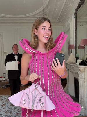 A woman in a bright pink dress holds a pink purse shaped like a jacket and smiles at the camera. A man in a suit stands in the background carrying a tray. The setting appears to be a well-lit room with elaborate architectural details.