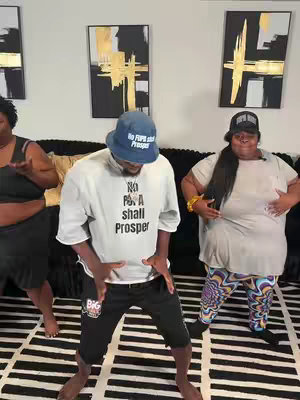 Three people dancing in a living room. A man in the center wearing black shorts, a white sweatshirt with the text “No P**** Shall Prosper”, and a blue hat. Two women flank him; one on the left wearing a black top and the other dancing on the right wearing a gray top and colorful patterned pants. There are framed art pieces on the wall behind them.