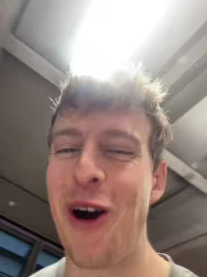 A selfie-style shot of a young man, slightly angled upwards, filling the frame. He has short, light-colored hair, and his facial expression suggests a mixture of surprise and amusement, with one eye partially closed and his mouth slightly open. Bright, diffused light emanates from above, casting a soft glow on his face and hair, which appears somewhat tousled. The background is indistinct, suggesting an indoor setting with a light fixture overhead, indicated by the bright illumination.