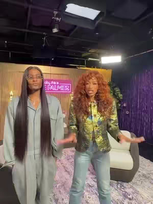 Two women stand side-by-side in a brightly lit setting. The woman on the left has long, straight, black hair and wears a light-colored pantsuit with glasses. The other woman, on the right, has voluminous auburn hair and wears a metallic blazer with a reflective sheen and light-colored jeans.
