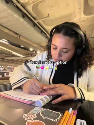 A person with curly hair writes in a notebook while wearing headphones. The person is seated at a desk or table, with a pen in hand, and there are some stationery items on the table with the notebook, including some colorful markers. There is a text overlay at the top of the screen that reads "Failure is not a option". Inside a library setting.