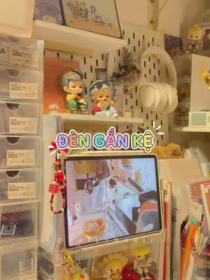 A well-lit photo of a study desk with various items. The desk features a tablet displaying a video, shelves filled with stationery and decorative items, and clear plastic storage containers. The overall aesthetic is neat and organized, with a soft, inviting atmosphere.