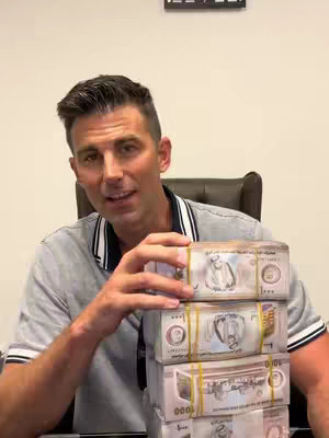 A man is seated in a chair, holding a stack of banded bundles of paper currency. The bundles appear to be currency, possibly from the United Arab Emirates, given the caption content and appearance. The man is looking at the camera, seemingly talking. The background appears to be an office-like setting with a framed item on the back wall.