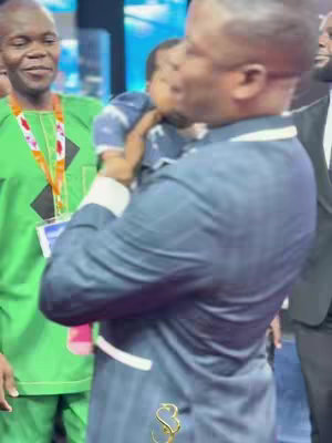 A man in a green shirt with a sash is smiling to the left. A man in a blue suit holds a baby in his arms, the baby is looking toward the right. A man on the right is wearing a black suit.
