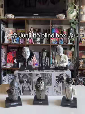 The cover image showcases a collection of Junji Ito manga character figurines displayed on a white table, with a shelf in the background filled with manga and other collectibles. Above the figurines is a shelf with decorative bowls and a television. The text overlayed reads "Junji Ito blind box". Overall, the composition has a clean, organized aesthetic, with a focus on the figurines.