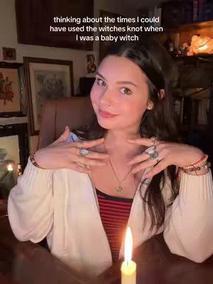 A woman with dark hair, fair skin, and a gentle smile is centered in the frame. She wears a white blouse over a red-striped top and various rings on her fingers. Her hands are positioned near her neck and face, and a lit candle is at the bottom, partially obscuring a wooden table.