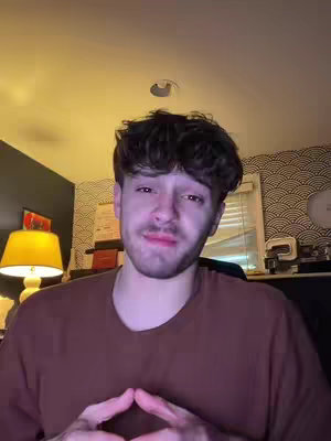 A man with dark curly hair is centered in a medium shot. He's wearing a dark brown shirt. His gaze is direct, and his hands are clasped together near his chest. The background has a variety of elements, including a desk lamp and a patterned wall. The lighting appears warm and natural.