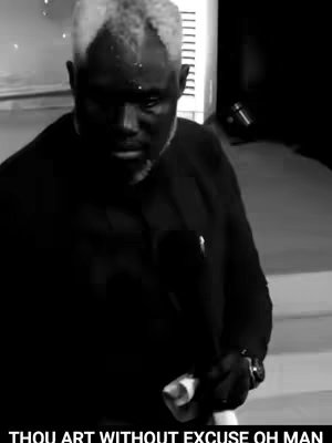 A black and white image features a man with short, gray-streaked hair and a serious expression. The man is wearing a dark-colored suit and is positioned in front of what appears to be a stage or podium. The lighting is dim, casting shadows that emphasize his facial features. Below his face, in a clear sans-serif font, the text 'THOU ART WITHOUT EXCUSE OH MAN' is displayed.