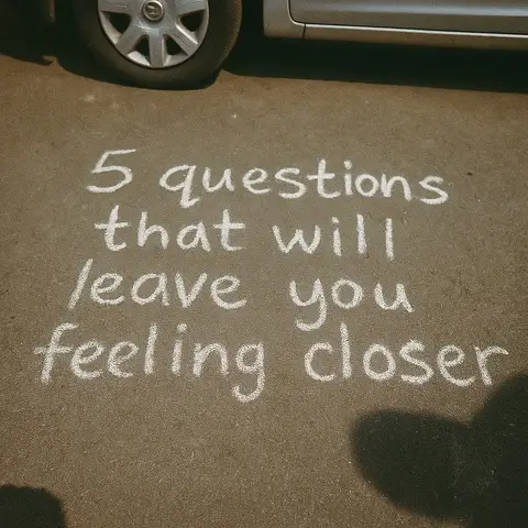 The image shows a section of a paved surface, possibly asphalt or concrete, with large white chalk lettering on it. The text reads, in a somewhat irregular font, '5 questions that will leave you feeling closer'. The letters are distinct, and the spacing is relatively neat. Above and to the left of the text, a portion of a car, including the tire and a section of the body, is visible, suggesting the setting is outdoors, likely a parking lot or driveway. The lighting appears to be natural, and the overall composition is simple and direct, focusing the viewer's attention on the message.