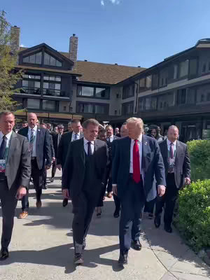 An outdoor shot features numerous men in suits walking towards the viewer. Prominent figures include men resembling Emmanuel Macron and Donald Trump, walking side by side at the center. A well-maintained building with a brown roof and a clear sky is visible in the background.